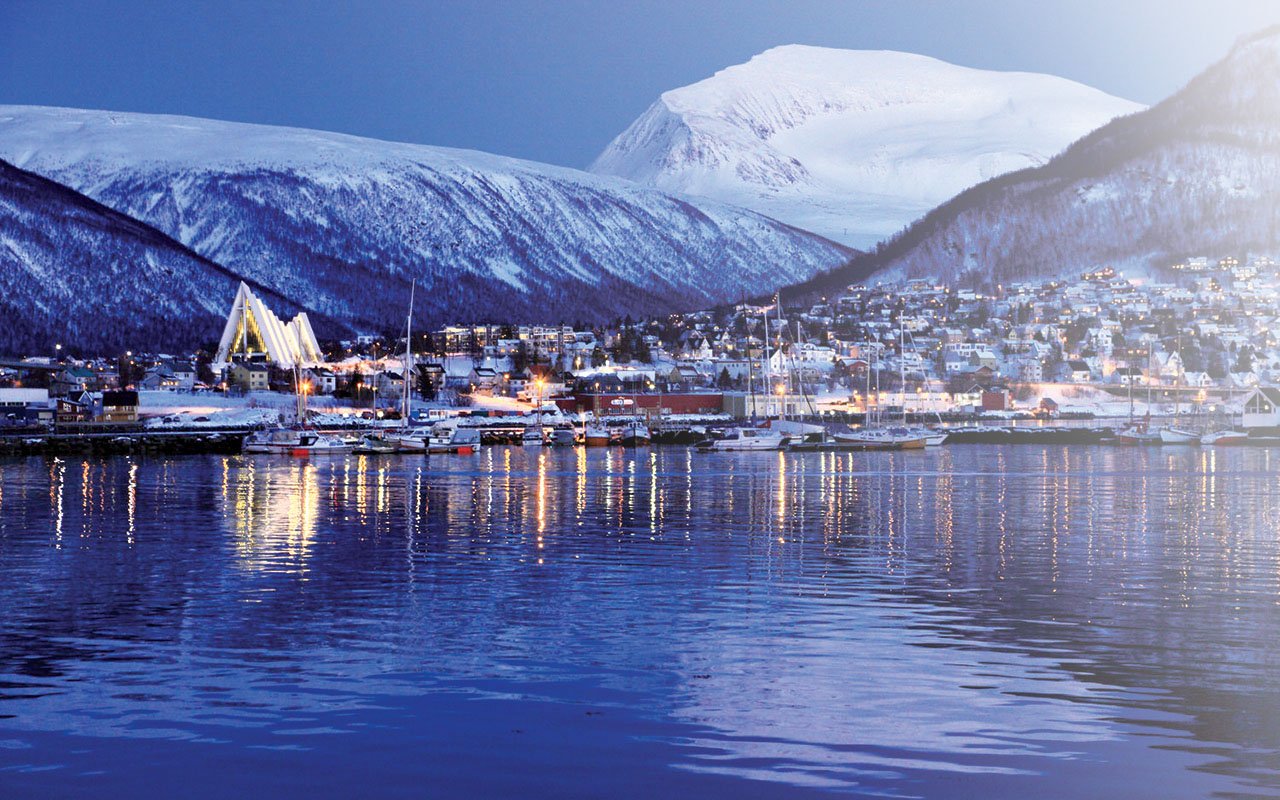Bilde av Tromsø fra vannet,i skomringen . Du kan se Ishavskatedralen og fjell med snø i bakgrunnen.