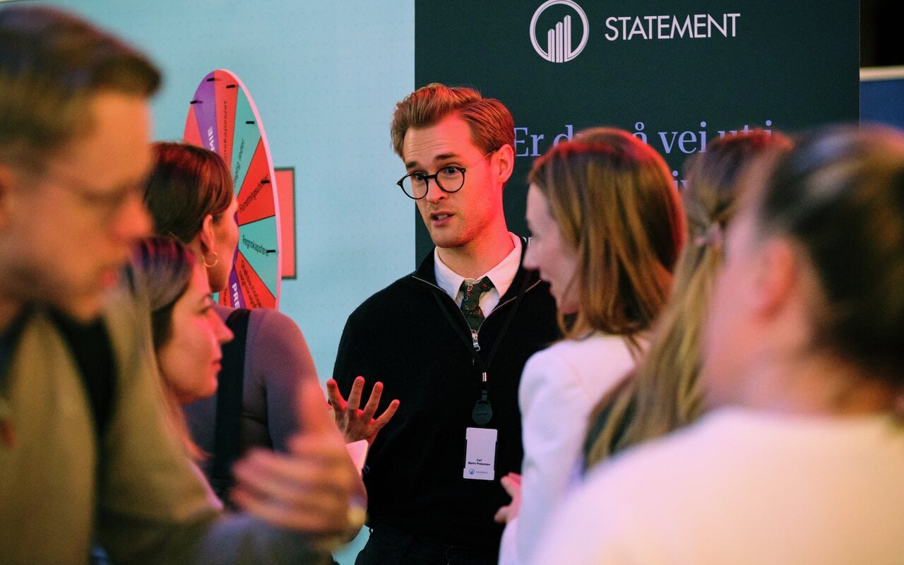 En gruppe mennesker står samlet på et arrangement og snakker sammen i et messeområde. I bakgrunnen er det en stand med teksten “STATEMENT” og et fargerikt lykkehjul. Personer i forgrunnen er i dialog, og en av dem bærer et navnekort rundt halsen.